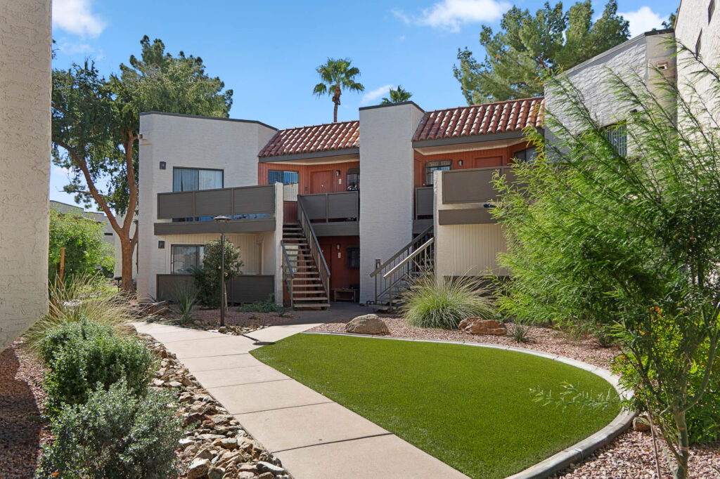 Modern white apartment building with two levels, exterior staircases, red accents, and a red-tiled roof. Landscaping includes rocks, bushes, and a curved path with artificial grass under a clear blue sky.