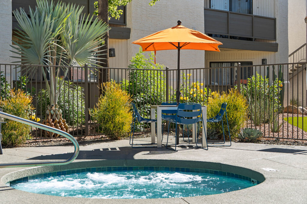 A round outdoor hot tub with bubbling water is in the foreground, surrounded by a patio area with blue chairs and a table under an orange umbrella, near plants and a fenced apartment building.
