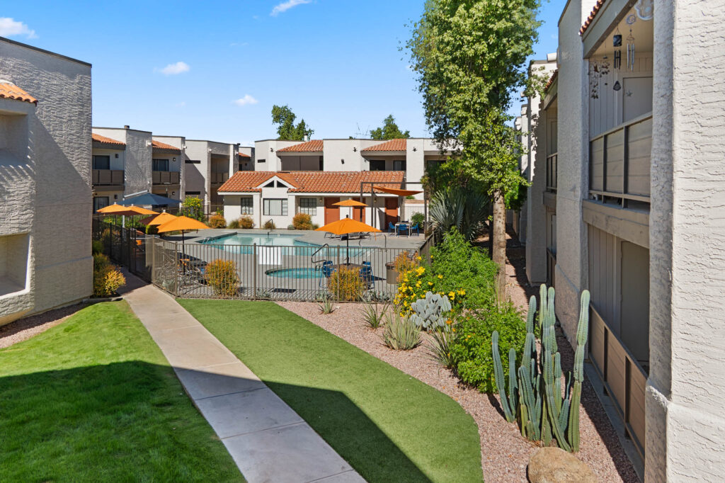 A view of an apartment complex courtyard featuring a fenced swimming pool with orange umbrellas, green plants, cacti, and well-maintained lawns, surrounded by beige buildings with red-tiled roofs under a blue sky.