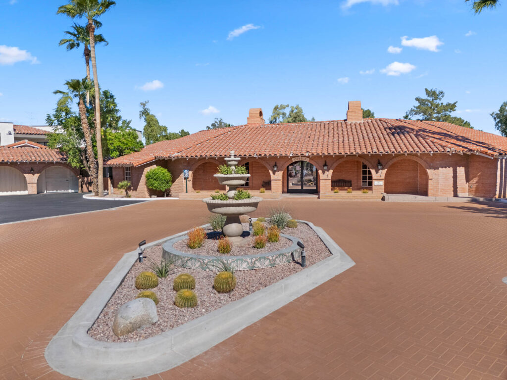 A large southwestern-style house with a red tile roof and brick exterior. A roundabout driveway surrounds a decorative stone fountain and desert landscaping with rocks, gravel, and cacti under a clear blue sky.