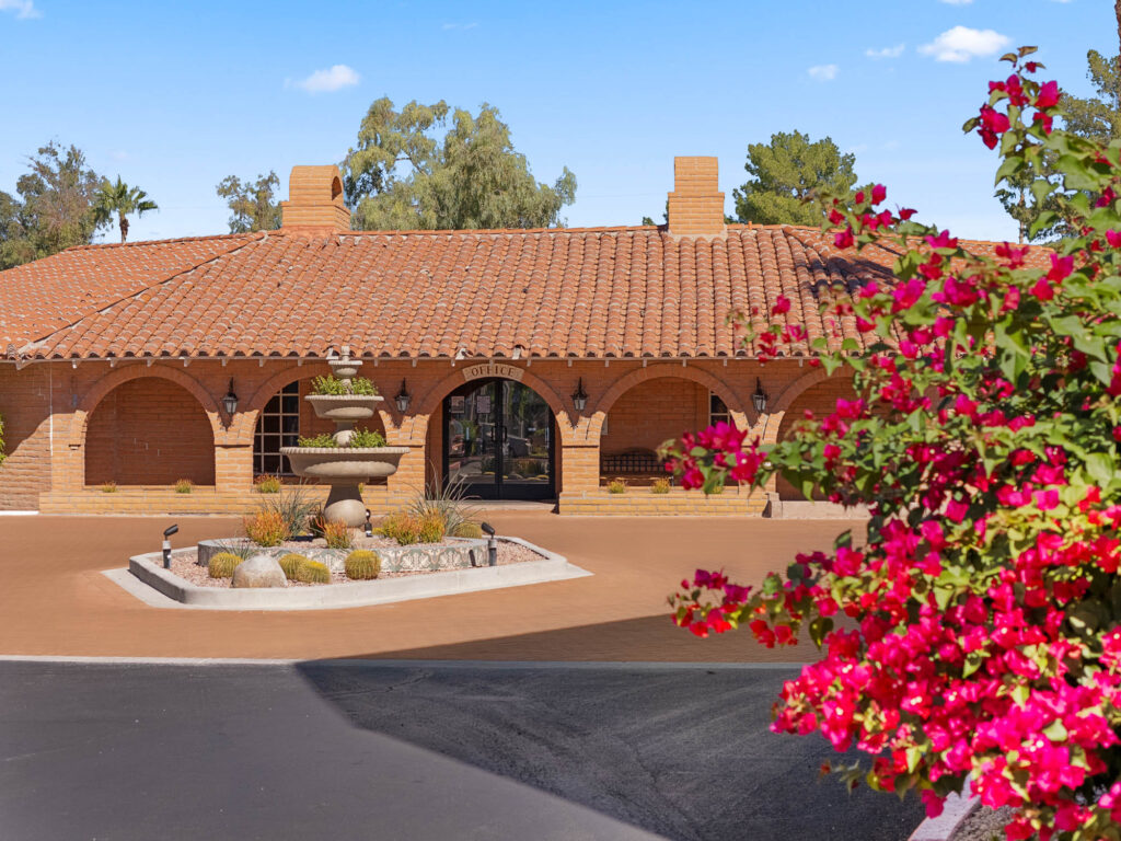 A single-story building with a red tiled roof and arched doorways, featuring a central stone fountain and surrounded by desert landscaping; bright pink flowers are in the foreground.