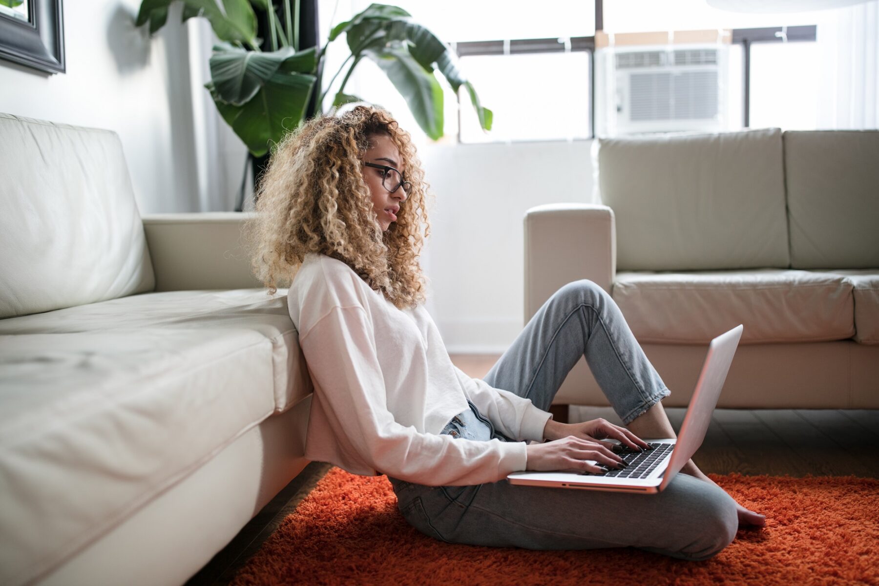 A person with curly hair and glasses sits on an orange rug, leaning against a white sofa while using a laptop in a bright living room with large windows and green plants.